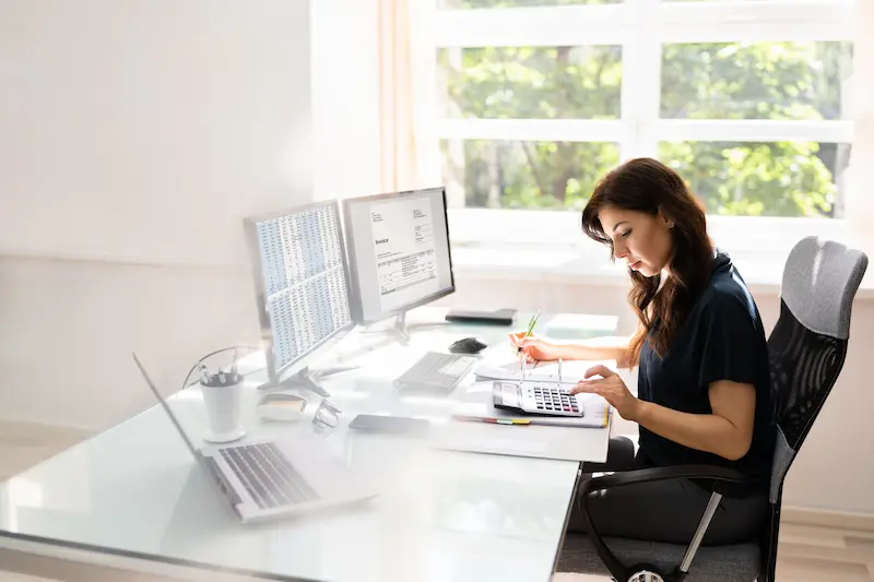 Eine junge Frau sitzt am Computer-Arbeitsplatz über Aktenordner und Taschenrechner gebeugt